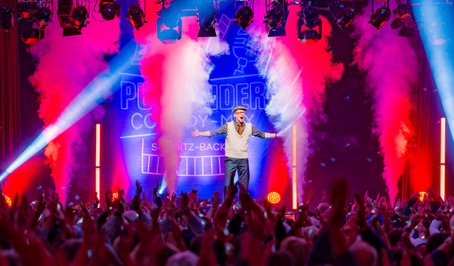 Schmitz-Backes auf der Bühne des Pullunder-Comedy-Mix vor jubelndem Publikum, Neon-Logo und Bühnennebel